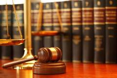 A gavel rests on sounding block next to a brass scale of justice in front of a long row of law books on a desk in a law office. Photographed with a very shallow depth of field.